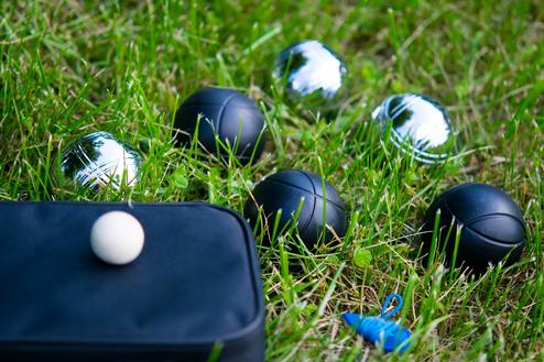 Schwarze Boulekugeln und eine weiße Kugel liegen auf grünem Gras neben einer kleinen, schwarzen Tasche.