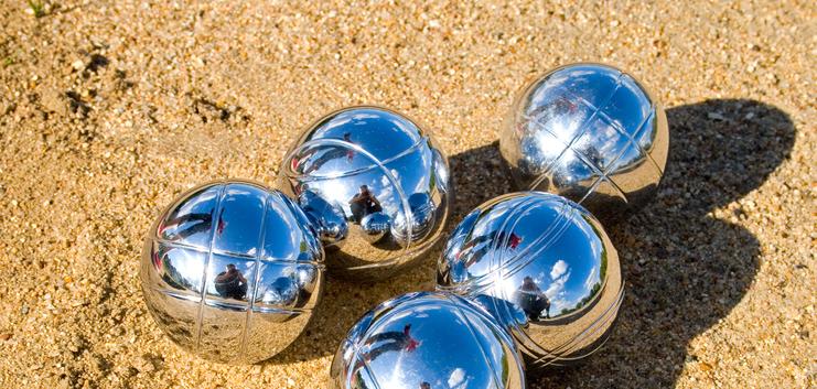 Fünf glänzende Boulekugeln liegen auf sandigem Untergrund, reflektieren Licht und Umgebung.