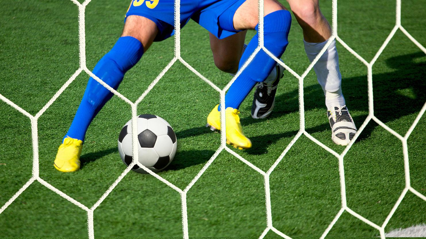 Zwei Fußballspieler in blauen und weißen Trikots kämpfen um einen Fußball vor einem Tor mit Netz.