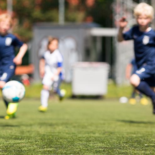 Zwei Kinder in Fußballtrikots rennen auf einem Kunstrasenplatz, ein Ball rollt in der Nähe.