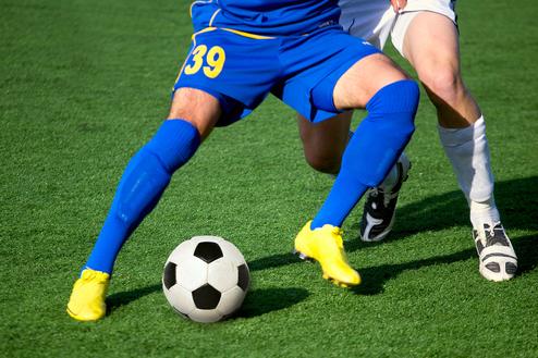 Zwei Fußballspieler kämpfen um den Ball auf einem grünen Kunstrasenplatz.