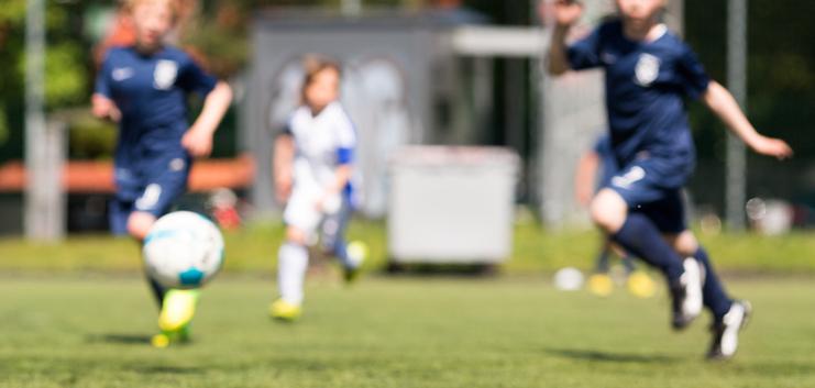 Zwei Kinder in Fußballtrikots rennen auf einem Kunstrasenplatz, ein Ball rollt in der Nähe.