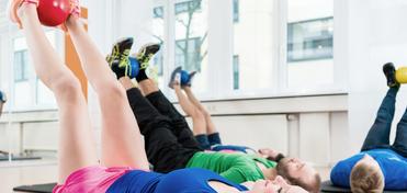 Gruppentraining: Frauen und Männer liegen auf Matten, heben Beine mit Medizinbällen in einem modernen Fitnessstudio.