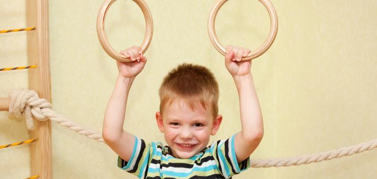 Junge mit einem gestreiften T-Shirt hält sich an zwei Holzringen in einem Spielraum fest und lächelt.