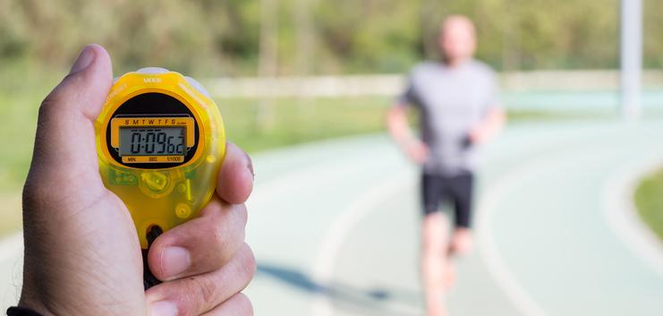 Hand hält eine gelbe Stoppuhr mit 00:08:66, während ein Jogger auf einer Laufbahn im Hintergrund läuft.
