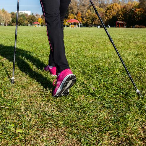 Frau in sportlicher Kleidung mit Nordic Walking-Stöcken läuft über eine grüne Wiese unter blauem Himmel.