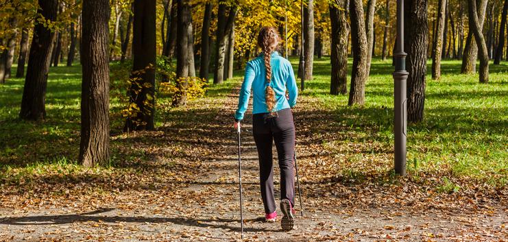 Frau mit langem Zopf geht mit Wanderstöcken einen herbstlichen Weg zwischen Bäumen, umgeben von buntem Laub.